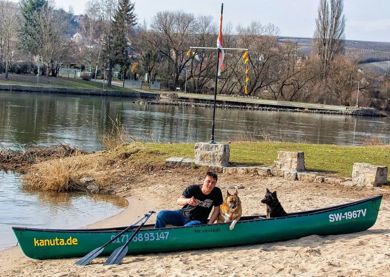 Ein Mann in einem grünen Kanu mit zwei Hunden im Sand.